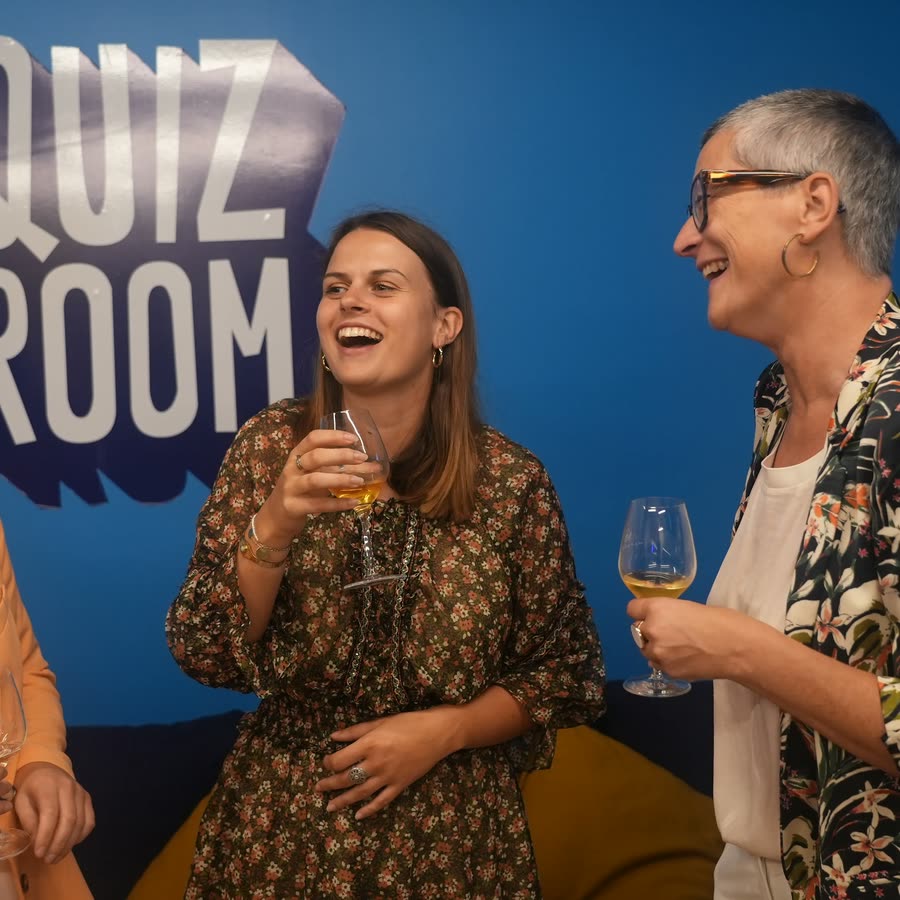 Trois joueuses riant, un verre à la main, devant le mur Quiz Room